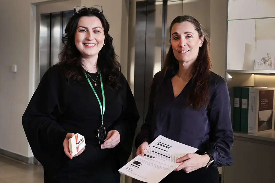 Cornelia och Malin står bredvid varandra i stashusets reception. De håller upp ett utskrivet maskerat dokument och verktyg som används för maskering på det traditionella sättet.