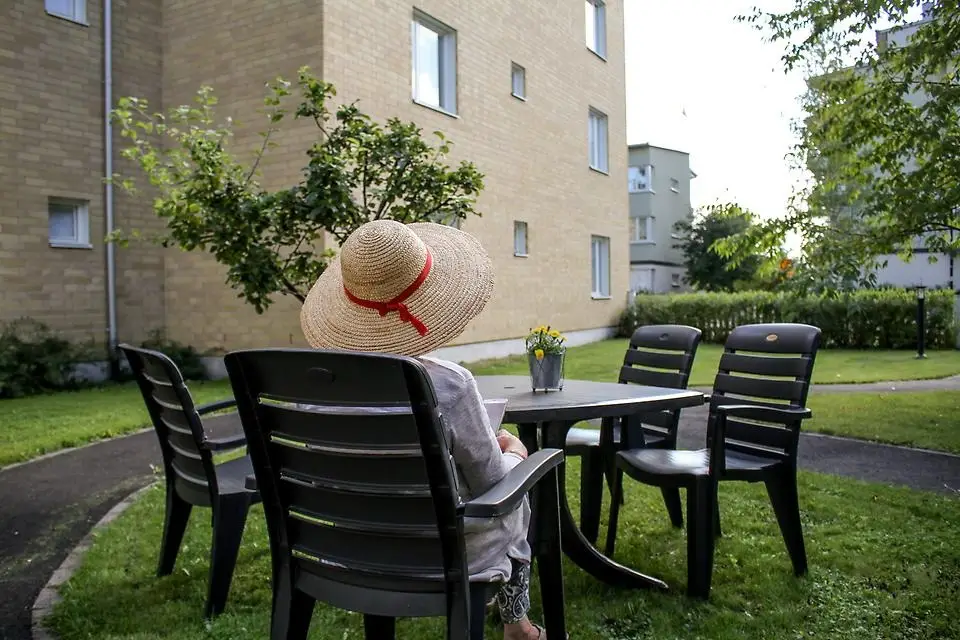 Person med sommarhatt sitter i trädgårdsmöbel på innergård vid flerfamiljshus om sommaren.